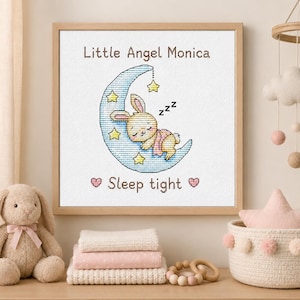 May include: A square framed cross-stitch embroidery featuring a sleeping bunny on a light blue crescent moon with yellow stars and the text Little Angel Monica and Sleep tight in brown lettering, accented by two small pink hearts on a white fabric.