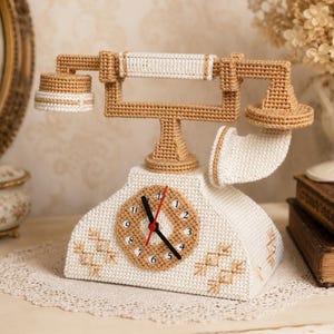 Può includere: Un orologio all'uncinetto a forma di telefono in filato color crema e marrone chiaro. La base presenta un quadrante circolare con lancette nere e una lancetta dei secondi rossa, arricchito da ricami geometrici color marrone chiaro.