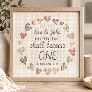 Puede incluir: Un bordado en punto de cruz en un marco cuadrado con una corona circular de corazones pequeños en tonos rosa, beige y marrón. El texto dice: 25.06.2026, Eve & John, And the two shall become ONE, EPHESIANS 5:31, centrado sobre tela blanca.