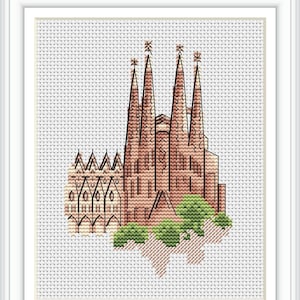 Puede incluir: Punto de cruz de la Sagrada Familia en Barcelona, España. La obra presenta las agujas y la fachada de la catedral en tonos marrones y beige, con arbustos verdes en la base. La pieza está enmarcada en un marco blanco.
