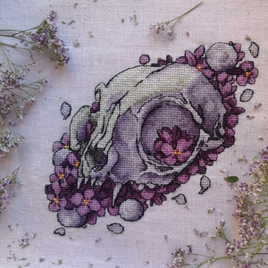 May include: Cross-stitch artwork depicting a skull design encircled by purple flowers. The skull is mainly white and grey, with purple shading. The flowers are a vibrant purple. The artwork is on a white fabric background.