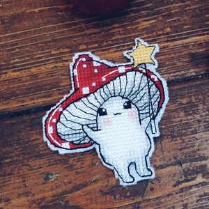 Könnte beinhalten: Ein Kreuzstich-Patch mit einem skurrilen Pilzcharakter mit roter und weiß gepunkteter Kappe. Der Pilz hält einen sternförmigen Zauberstab. Der Patch ist weiß mit schwarzen Umrissen und vor einem Holzgrund mit zwei roten Äpfeln.