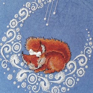 May include: Cross-stitch art featuring a sleeping fox on a crescent moon. The fox is orange with a white bow, resting on a white embroidered moon against a blue backdrop. The artwork includes small white embellishments and a "handmade" tag.