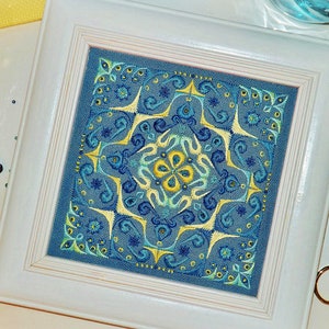May include: A framed, square embroidery with a detailed, symmetrical design in shades of blue and yellow. The artwork is set in a white wooden frame. Decorative items like scissors, beads, and a small bottle are arranged around the frame.