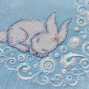 May include: A cross-stitch design featuring a sleeping bunny with a small flower on its head. The bunny is white and lavender, set against a light blue background. Delicate white and light blue swirls and floral accents surround the bunny, creating a whimsical scene.
