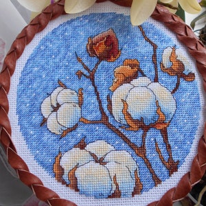 May include: A cross-stitch embroidery of cotton plants with white fluffy bolls and brown stems against a blue background. The embroidery is framed by a brown braided leather border. Cream-colored orchids with purple and pink accents are in the background.