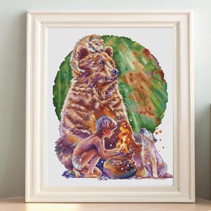 May include: Framed cross-stitch art depicting a bear with a cub on its back, a child, and a wolf around a fire. The artwork features a green and brown background, with a light-colored frame. The scene is set on a wooden table.