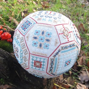 May include: A decorative sphere with detailed cross-stitch patterns in blue, red, and tan. The sphere is adorned with small beads and sits on a weathered wooden surface, surrounded by greenery and berries. The sphere's design is geometric.