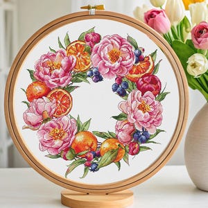 May include: A cross-stitch embroidery hoop featuring a floral wreath design. The wreath includes pink peonies, orange slices, blueberries, and green leaves. The embroidery is set in a wooden hoop and displayed on a wooden stand.