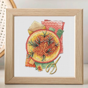 Puede incluir: Una obra de arte de punto de cruz enmarcada que presenta una representación detallada de una fruta, posiblemente una granada, con una combinación de colores amarillo y naranja. La obra de arte está acompañada de retazos de tela y tijeras doradas.