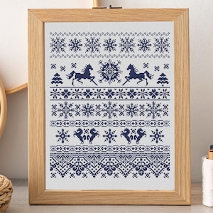 May include: A framed cross-stitch with a navy blue and white winter pattern. The design includes snowflakes, horses, and geometric shapes. The wooden frame holds the artwork, displayed on a light-coloured surface.