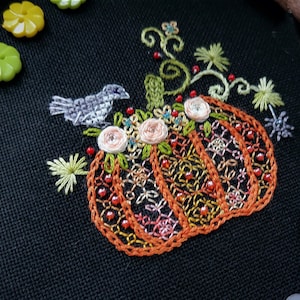 May include: Embroidered pumpkin design on black fabric. The pumpkin is orange with intricate stitching and bead details. Features a small gray bird, pink flowers, and green leaves. Includes a wooden embroidery hoop and orange thread.
