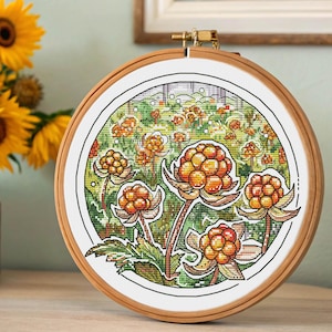 May include: A cross-stitch embroidery in a wooden hoop features cloudberries with orange and yellow berries and green leaves. The background shows a field of cloudberries. A vase of sunflowers sits to the left of the embroidery.