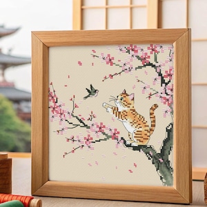 Puede incluir: Arte de punto de cruz en un marco de madera que representa un gato naranja extendiendo la pata hacia un pájaro en una rama floreciente. La obra presenta flores rosas y un fondo beige, con una pagoda japonesa al fondo.