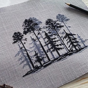 May include: Cross-stitch embroidery of a forest scene with tall pine trees in black and blue hues. The artwork is on light grey linen fabric. A pencil and sharpener are visible in the background.