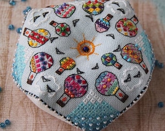 May include: A light blue, pentagon-shaped, embroidered pincushion with a sunburst design and colorful hot air balloon motifs. The edges are trimmed with blue beads. Scattered blue beads surround the pincushion.