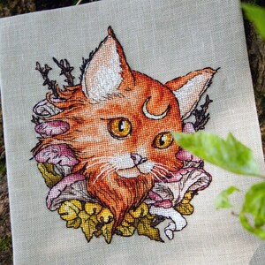 Könnte beinhalten: Kreuzstich-Kunstwerk mit einem stilisierten orangefarbenen Fuchs mit gelben Augen, einem Mond und einem Bart. Der Fuchs ist von Pilzen und Blättern umgeben, gestickt auf hellem beigen Stoff. Das Kunstwerk wird vor einem Baumstamm präsentiert.