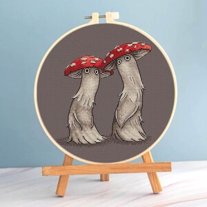May include: Cross-stitch embroidery of two whimsical mushrooms with red and white spotted caps, gray stems, and expressive eyes. The artwork is displayed in a wooden embroidery hoop, mounted on a small wooden easel, set against a blue background.