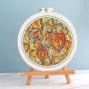 May include: A completed cross-stitch embroidery in a round hoop, featuring a detailed botanical design of orange and yellow Chinese lantern plants. The artwork is displayed on a small wooden easel against a light blue background.