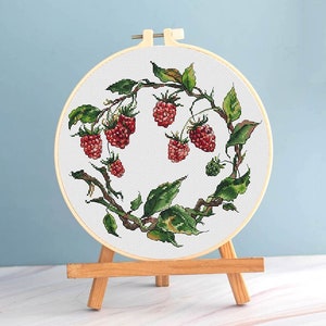 May include: A cross-stitch embroidery hoop featuring a detailed design of red raspberries and green leaves on a white background. The embroidery is set in a wooden hoop and displayed on a small wooden easel.