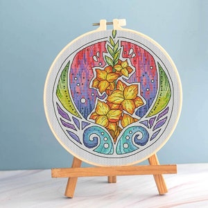 May include: A completed cross-stitch embroidery in a wooden hoop. The design features yellow flowers with green leaves, set against a colorful background of red, purple, and blue. The embroidery is displayed on a small wooden easel.