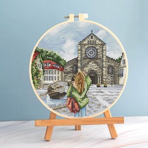 Peut inclure: Une broderie au point de croix représentant une scène de ville européenne, avec une grande église en pierre et une personne aux cheveux blonds. La broderie est présentée dans un cercle en bois et posée sur un petit chevalet en bois.