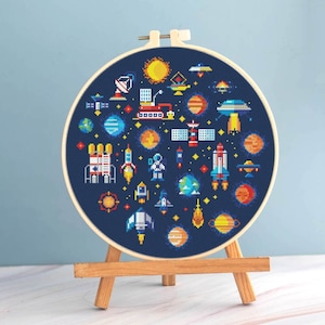 May include: A cross-stitch embroidery hoop featuring a colorful space-themed design. The artwork includes planets, rockets, satellites, and a sun, all set against a dark blue background. The hoop is displayed on a small wooden easel.