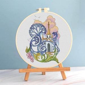 Può includere: Un telaio da ricamo a punto croce con un castello, un albero e elementi floreali. L'opera d'arte utilizza sfumature di blu, verde e rosa. Il ricamo è esposto su un cavalletto di legno su sfondo azzurro.