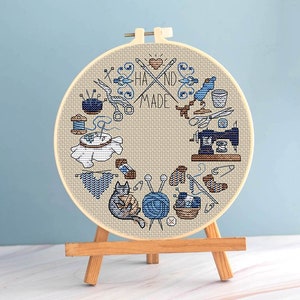 May include: A cross-stitch embroidery hoop with a beige fabric background. The design features blue illustrations of sewing tools, yarn, and the words "HAND MADE". The hoop is displayed on a small wooden easel.