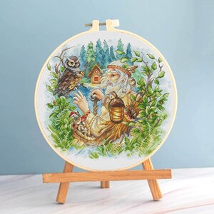 Könnte beinhalten: Kreuzstich-Stickerei in einem Holzrahmen, die einen alten Mann mit langem, weißem Bart, eine Eule und eine kleine Hütte in einer Waldszene zeigt. Das Kunstwerk weist detaillierte Stiche mit leuchtenden Farben auf, vor einem hellblauen Hintergrund, auf einer kleinen Holzstaffelei.