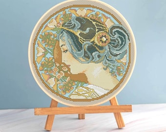 Pode incluir: Uma obra de arte de ponto cruz concluída em um bastidor de madeira, apresentando o perfil de uma mulher com cabelos esvoaçantes e detalhes florais. A paleta de cores inclui tons de azul, dourado e bege. A obra de arte é exibida em um pequeno cavalete de madeira.