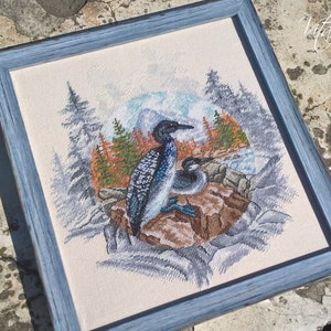 May include: Cross-stitch artwork of two loons on a rocky shore, framed in a light blue wooden frame. The scene includes a backdrop of evergreen trees, mountains, and a body of water. The artwork is detailed with various colors.
