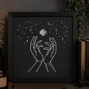 Può includere: Un'opera d'arte a punto croce in bianco e nero incorniciata raffigura due mani che si protendono verso una luna e delle stelle. L'opera è esposta su una superficie di legno con una candela accesa e una lampada decorativa.