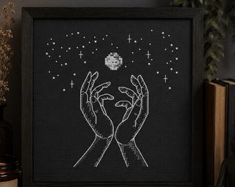 May include: A framed black and white cross-stitch art piece depicts two hands reaching up towards a moon and stars. The artwork is displayed on a wooden surface with a lit candle and a decorative lamp.