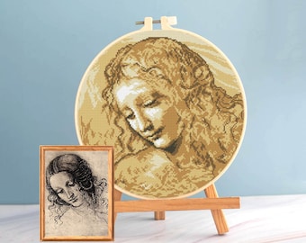 Pode incluir: Uma bordado de ponto cruz de um retrato de mulher num círculo de madeira, exposto num pequeno cavalete de madeira. Uma impressão emoldurada do mesmo retrato está ao lado. A obra de arte apresenta uma paleta de cores sépia.