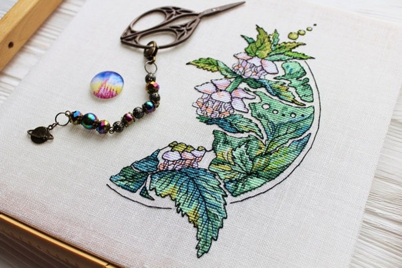 May include: Cross-stitch embroidery of a floral design with green leaves and pink flowers. A pair of embroidery scissors, a decorative charm bracelet with iridescent beads, and a small, colorful button are also present.
