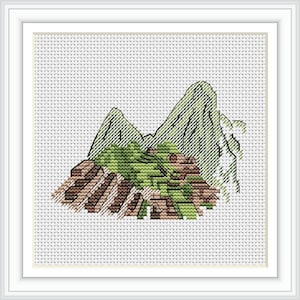 Può includere: Punto croce di un paesaggio montano con cime verdi, strutture marroni e fogliame verde. L'opera è incorniciata in una cornice quadrata bianca, su uno sfondo bianco con un motivo a punti.