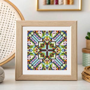 May include: A framed cross-stitch artwork featuring a geometric, symmetrical design. The design incorporates a variety of colors, including blue, green, orange, and brown, creating a vibrant and intricate pattern. The artwork is displayed in a light-colored wooden frame.