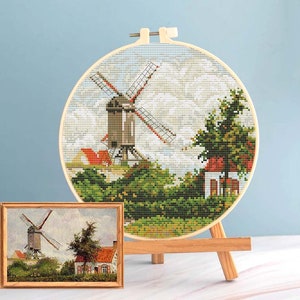 May include: A cross-stitch embroidery of a windmill scene, displayed in a wooden hoop on a small easel. The scene includes a windmill, houses with red roofs, and green foliage. A framed painting of the same scene is also present.