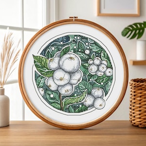 Könnte beinhalten: Eine Kreuzstich-Stickerei mit weißen Beeren und grünen Blättern, die in einem Holzreifen eingefasst ist. Das Design weist detaillierte Stiche und ein botanisches Thema auf, ideal für die Inneneinrichtung. Das Kunstwerk wird auf einer Holzoberfläche präsentiert.