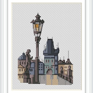 May include: Cross-stitch artwork of a European cityscape featuring a tall lamppost, a statue, and various buildings. The buildings have blue, tan, and gray tones. The artwork is framed in a white border.