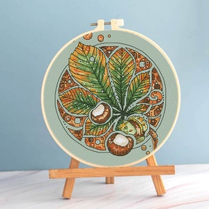 May include: A cross-stitch embroidery of a chestnut leaf and nuts, displayed in a wooden hoop. The leaf features green, yellow, and orange hues, with brown nuts. The embroidery is set against a light blue background and displayed on a small wooden easel.