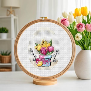 Könnte beinhalten: Ein Kreuzstich-Stickrahmen mit einem frühlingshaften Blumendesign. Das Design zeigt gelbe und rosa Tulpen in einem blau-rosa Topf, mit zwei verzierten Ostereiern. Der Rahmen steht auf einem Holzständer.
