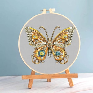 Könnte beinhalten: Eine Kreuzstich-Stickerei eines Schmetterlings mit Steampunk-Design. Der Schmetterling hat komplizierte Details in Gold-, Braun- und Türkistönen, vor einem grauen Hintergrund in einem hölzernen Stickrahmen. Der Rahmen wird auf einer kleinen Holzstaffelei ausgestellt.