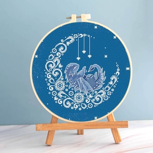 May include: A cross-stitch embroidery hoop featuring a blue dragon nestled in a crescent moon design. The dragon and moon are white, with intricate floral and star details. The embroidery is displayed on a small wooden easel.