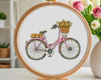 May include: A cross-stitch embroidery of a pink bicycle with a basket of flowers. The bicycle has yellow wheels and a brown seat. The embroidery is displayed in a round wooden hoop, set on a wooden stand.