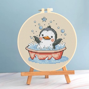 Puede incluir: Un bastidor de bordado de punto de cruz con un pingüino de dibujos animados bañándose en una bañera roja. El pingüino es negro, blanco y naranja, rodeado de burbujas y agua azules. Un pajarito está encima del pingüino. El bastidor está sobre un caballete de madera.