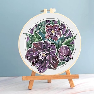 May include: A completed cross-stitch embroidery of purple flowers and green leaves, set in a round wooden hoop. The floral design is displayed on a small wooden easel against a light blue background. The embroidery features detailed stitching.