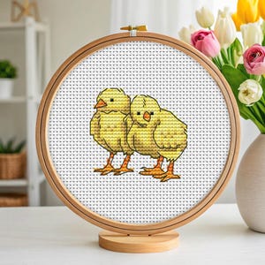 Könnte beinhalten: Ein Kreuzstich-Stickrahmen mit zwei gelben Küken. Die Küken haben orangefarbene Schnäbel und Füße und sind vor einem weißen Hintergrund platziert. Der Holzrahmen wird auf einem Holzständer präsentiert, mit einer Vase mit Blumen im Hintergrund.