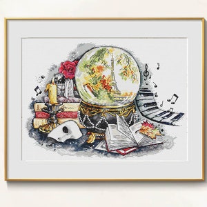 May include: Cross-stitch art print featuring a snow globe with the Eiffel Tower, books, a candle, a rose, and musical notes. The globe sits on a decorative base with a pearl necklace. A Phantom of the Opera mask is also present.